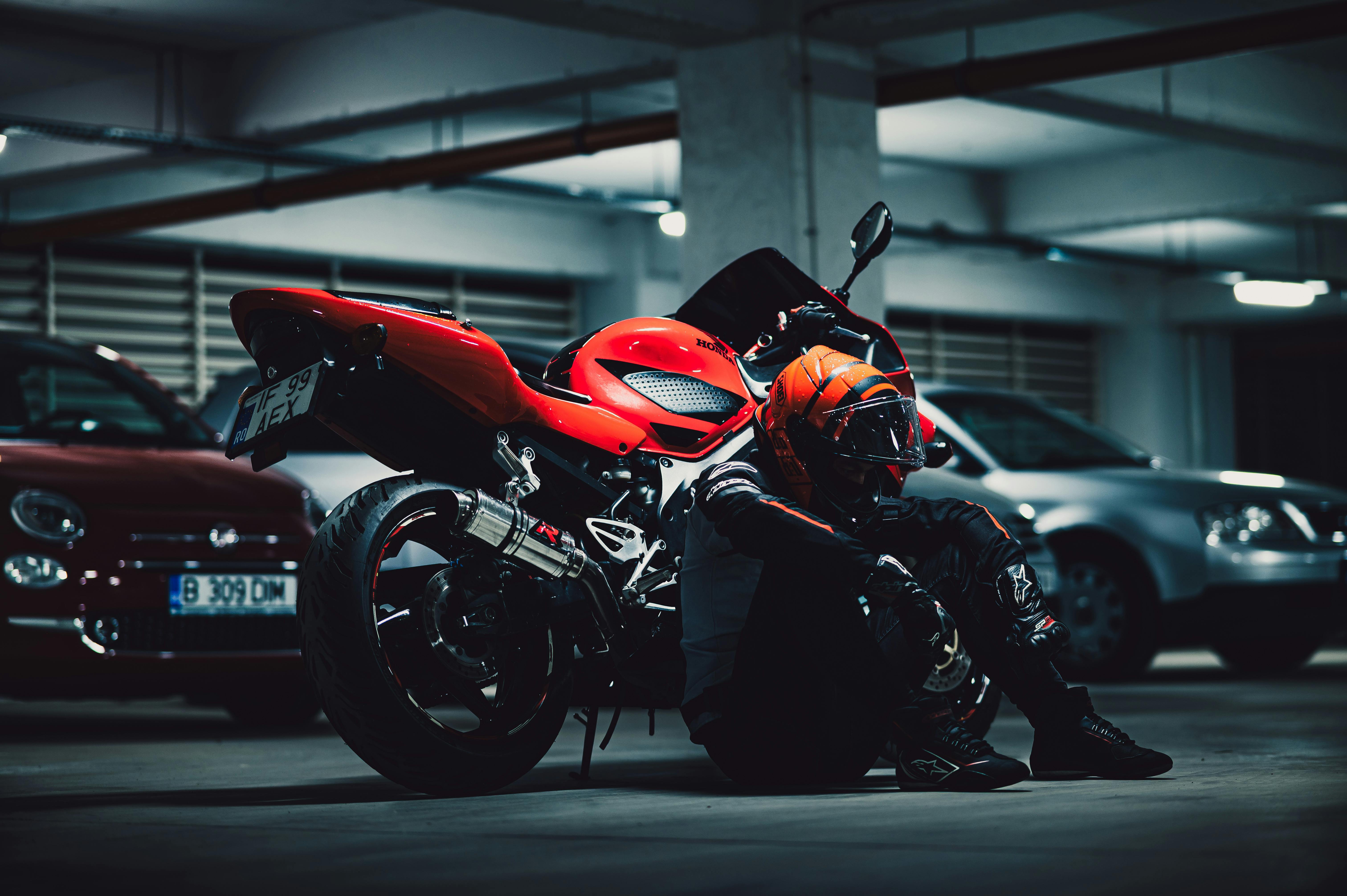 Motorcyclist with helmet sits beside red motorcycle in dimly lit parking garage.