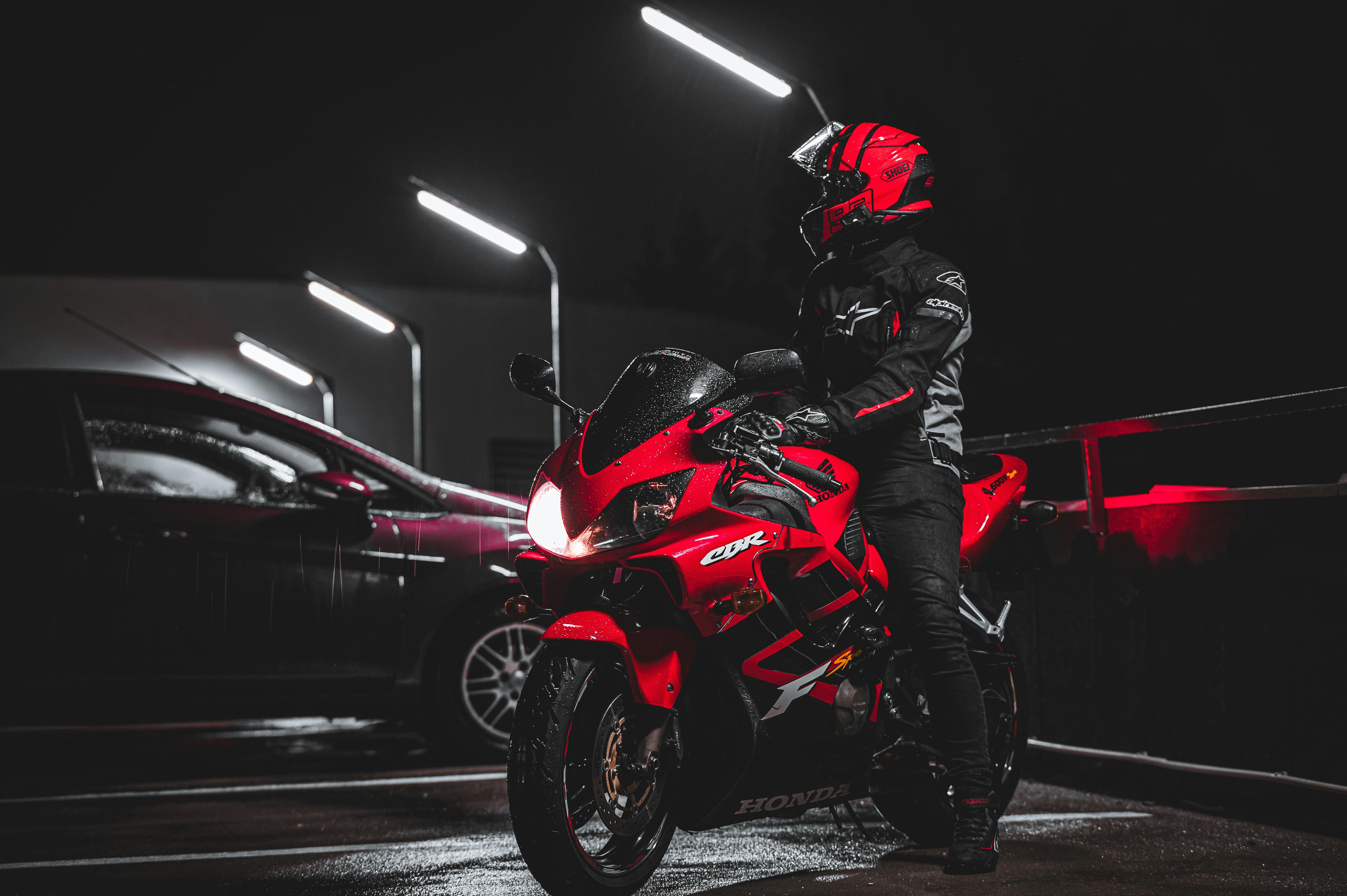 Motorcyclist in red gear standing beside red sports motorcycle at night in dimly lit parking area.