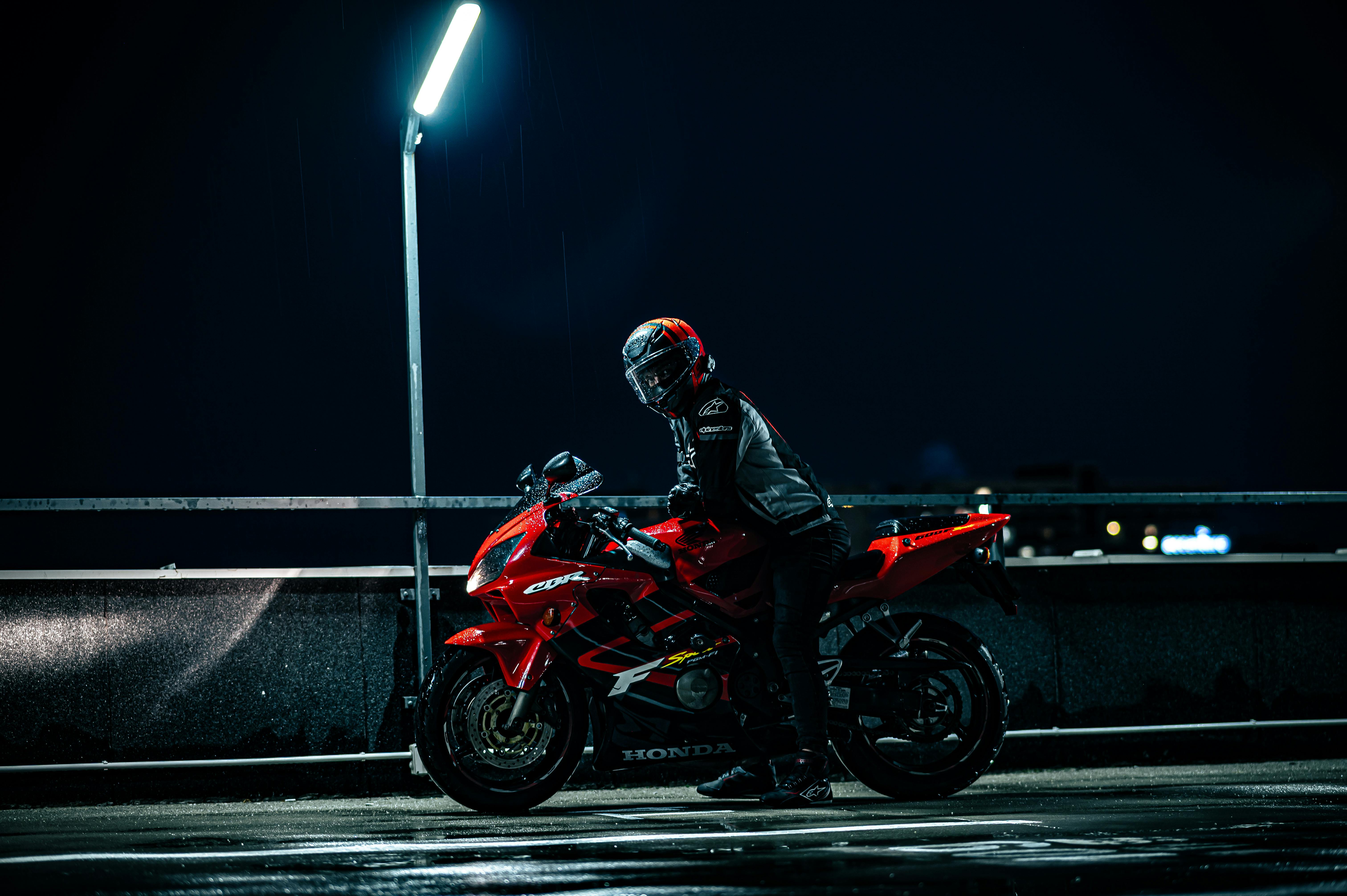 A motorcyclist in gear on a red sports bike under streetlight at night.